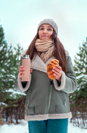 Portrait of a beautiful young woman with a flask and a croissant in the winter forestの写真素材