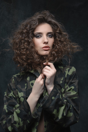 young girl closes from the cold in military uniform. curly brunette with blue eyes, stands on a dark backgroundの写真素材
