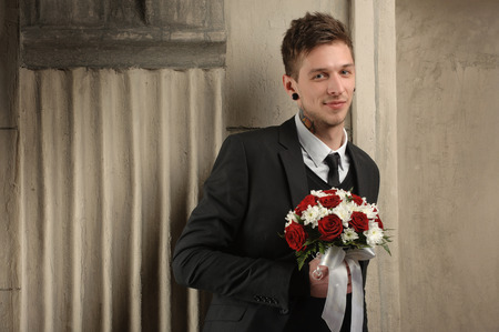 young stylish groom with tattoos waiting for his bride. in the hands holding flowers, leans against the wallの写真素材
