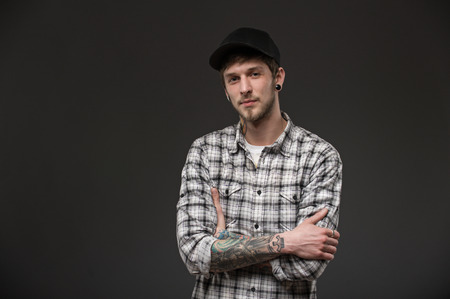 guy holds his hands together on his chest. dressed in a plaid shirt and a black cap. his body has tattoos and piercings in the earsの写真素材