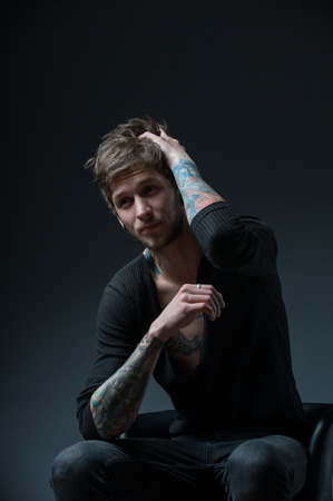 guy with messy hair sitting on a chair. With tattoos on both arms and piercings in the ear, on a black backgroundの写真素材