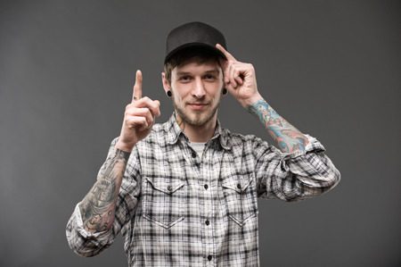 I have one idea. young guy shows his index finger upwards, the other hand holding the cap. He has a piercing in his ears and a tattoo on the bodyの写真素材