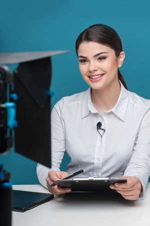 Waist up portrait of elegant woman reporter, who is smiling and looking at the camera, which is visual on the foreground, while sitting at the table and holding the folder and pen, isolated on a blue backgroundの写真素材
