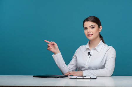 Waist up portrait of elegant woman reporter, who is looking at camera seriously and pointing her finger on the right hand aside while sitting at the table. She has long brown hair and a white blouse on which a little microphone is situated, isolated on a の写真素材