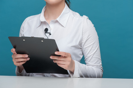 Close up portrait of elegant woman reporter, who is holding the folder in her hands and sitting at the table, isolated on a blue backgroundの写真素材