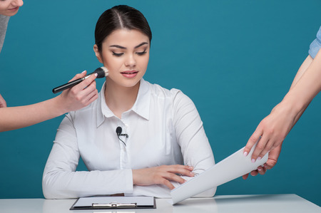 Waist up portrait of beautiful woman reporter with brown hair, who is sitting at the table while looking at the documents, which someone holds in their arms and another woman is doing make up for the newscaster, isolated on a blue backgroundの写真素材