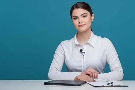 Waist up portrait of pretty woman reporter with brown hair, who is telling news and looking seriously at the camera, sitting at the table on which there is notebook folder and pen, isolated on a blue background and there is copy place in the left sideの写真素材
