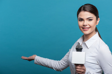 Half length portrait of  woman  reporter with long brown hair, who interviews and is smiling and looking at the camera holding the microphone. The girl shows with her right hand aside, isolated on a blue background and there is copy place in the left sideの写真素材