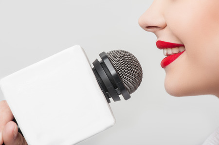 Close up portrait of beautiful woman reporter with Caucasian appearance, who is smiling and talking into the microphone while holding it with her right hand. She has bright red lips, isolated on a grey backgroundの写真素材