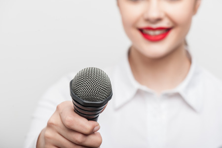 Close up portrait of beautiful woman tv reporter with Caucasian appearance and bright red lips, who is smiling and proposes someone to give her the interview holding the microphone, which is visible on the foreground while the portrait of the girl is sligの写真素材