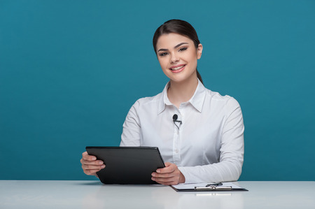 Waist up portrait of elegant woman reporter with Caucasian appearance, who is telling news and smiling and looking at the camera, sitting at the table and holding the notebook in her arms, isolated on a blue background and there is copy place in the left の写真素材