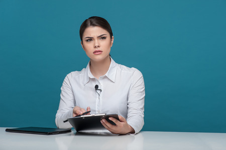 Waist up portrait of elegant woman reporter of Caucasian appearance, who is looking at the camera questioningly and trying to understand something holding the folder and pen in her arms and sitting at the table, isolated on a blue background and there is の写真素材