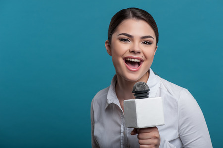 Waist up portrait of elegant young woman reporter, who interviews and is smiling at the camera speaking about something very emotionally and holding the microphone. She has Caucasian appearance, isolated on a blue background and there is copy place in theの写真素材