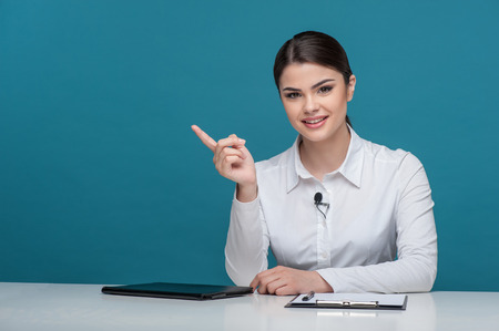 Waist up portrait of elegant woman reporter with brown hair, who is smiling and looking at the camera while sitting at the table and pointing with her finger aside, isolated on a blue background and there is copy place in the left sideの写真素材
