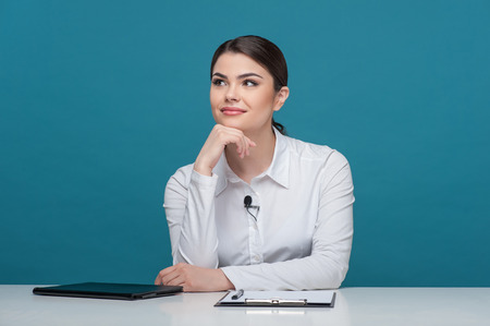 Waist up portrait of elegant woman reporter with Caucasian appearance, who is smiling and dreaming about something while sitting at the table and propping up her head with her right hand and her eyes are looking up, isolated on a blue background and thereの写真素材