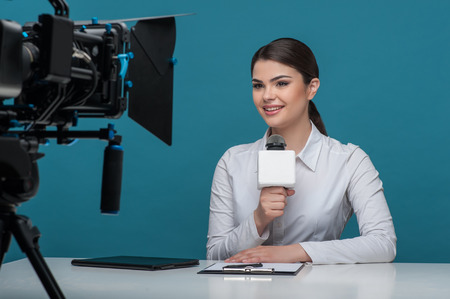 Waist up portrait of elegant woman reporter with brown hair, who is telling the news and smiling looking at the camera while sitting at the desk and holding the microphone, the camera is visual on a foreground, isolated on a blue backgroundの写真素材