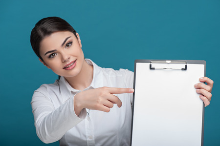 Waist up portrait of beautiful woman reporter, who is smiling and looking at the camera while holding the folder and pointing with her finger at it sitting at the, she has long brown hair, isolated on a blue backgroundの写真素材