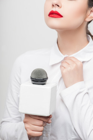 Close up portrait of beautiful woman reporter with brown hair, who is straightening the collar of her white blouse with her left hand nervously while another hand is holding the microphone. She has bright red lips, isolated on a grey backgroundの写真素材