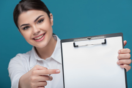 Waist up portrait of pretty woman reporter with Caucasian appearance, who is holding the folder and pointing her finger at it while smiling and looking at the camera and sitting at the table. The folder is visible on the foreground, isolated on a blue bacの写真素材