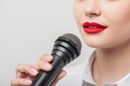 Close up portrait of beautiful woman reporter, who is holding the microphone and talking into it slightly smiling. She has Caucasian appearance and bright red lips, isolated on a grey background and there is copy place in the left sideの写真素材