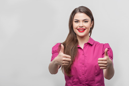 Everything is okay. Waist up portrait of happy beautiful young woman reporter with Caucasian appearance and red lips, who is smiling and raising her thumbs up as if everything were fine, isolated on a grey background and there is copy place in the left siの写真素材