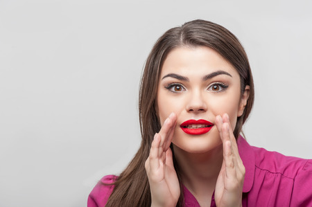 Waist up portrait of beautiful young woman reporter with brown hair and bright red lips, who is very surprised about something while raising up her hands near her mouth and looking at the camera, isolated on a grey background and there is copy place in thの写真素材