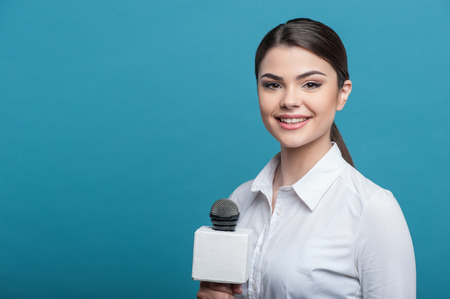 Waist up portrait of elegant woman reporter, who interviews and is smiling and looking at the camera holding the microphone. She has long brown hair and a white blouse, isolated on a blue background and there is copy place in the left sideの写真素材
