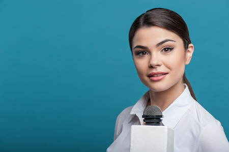 Waist up portrait of elegant woman reporter with Caucasian appearance, who is holding the microphone looking attentively at the camera and waiting for the answer. She has long brown hair and a white blouse, isolated on a blue background and there is copy の写真素材