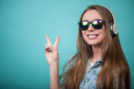 Waist up portrait of pretty hipster girl with headphones and sunglasses, who is listening to music, makes a gesture that symbolizes peace and smiling expressing joy, isolated on a blue background and there is cope place in the left sideの写真素材