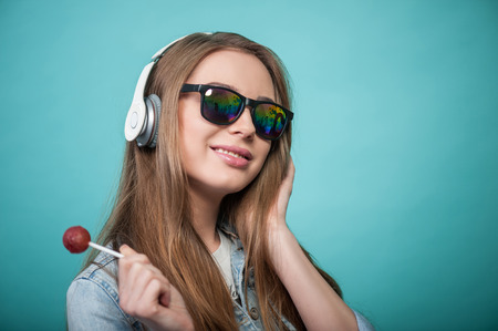 Waist up portrait of beautiful hipster girl with Caucasian appearance, who is holding a Lollipop and enjoining listening to music in headphones while touching it and smiling, isolated on a blue background and there is cope place in the right sideの写真素材