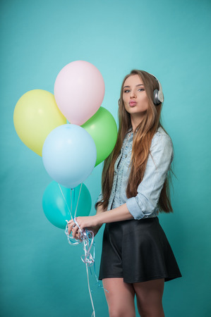 Full length portrait of beautiful hipster girl with Caucasian appearance, who is standing while holding varicolored balloons in her hand, wearing headphones listening to music looking at the camera mysteriously and sending blow kiss, isolated on blue backの写真素材