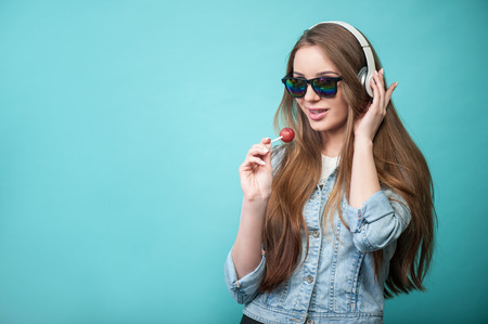 Waist up portrait of pretty hipster girl with Lollipop and glasses, who is looking mysteriously at the camera smiling and singing lovely listening to music, while touching the headphones which are on her head, isolated on a blue background and there is coの写真素材