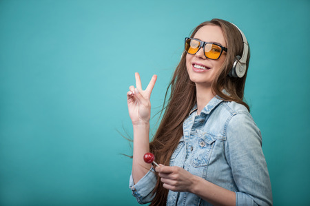 Waist up portrait of beautiful hipster girl with lollipop and sunglasses, who is listening to music in headphones and smiling looking at the camera, showing with her hand a gesture that symbolizes peace and positive mood, isolated on a blue background andの写真素材