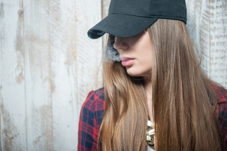 Waist up portrait of beautiful hipster girl with cap on her head, which closes her eyes so we can see only her mouth from which the smoke is coming. Girl is cad and depressed, isolated on a white background and there is copy place in the left sideの写真素材