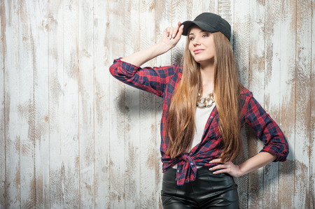 Waist up portrait of cheerful hipster girl with Caucasian appearance, who is standing near the white wall and looking towards mysteriously slightly smiling, touching her cap which is on her head, isolated on a white background and there is copy place in tの写真素材