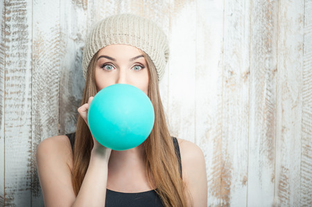 Pretty hipster woman is inflating colored balloonWaist up portrait of cheerful hipster girl with Caucasian appearance, who is standing near the white wall and is blowing balloon diligently and looking at the camera amazedly, isolated on a white backgroundの写真素材