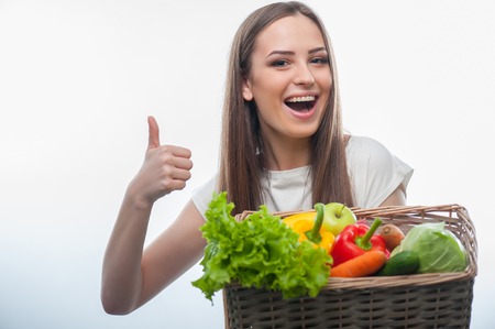 pretty girl happily holding the basket of healthy food. She is smiling and giving her thumbs up, isolated on a white backgroundの写真素材