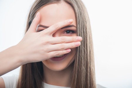 Close up portrait of cheerful girl showing happiness while smiling. She closes her face by her hand so that she is piping through it, isolated on a white backgroundの写真素材