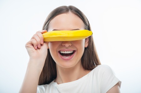 Cheerful girl is holding healthy banana playfully. She is close her eyes by it. Isolated on a white backgroundの写真素材