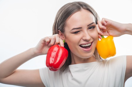 Beautiful girl winking at the camera. She is holding two peppers and raising them to her ears laughing and joking, isolated on a white backgroundの写真素材
