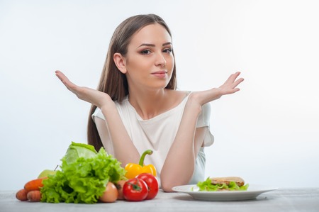 Beautiful girl cannot choose between healthy and unhealthy food. She gesturing and looking at the camera confused. Isolated on a white backgroundの写真素材