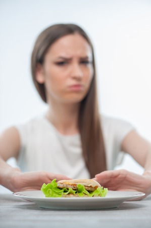 Pretty healthy woman is pushing aside the plate with harmful hamburger. Focus on hamburger, Isolated on a white backgroundの写真素材