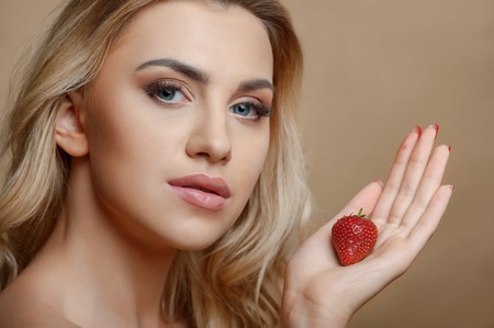 Beautiful girl is holding strawberry in her palm and showing it to camera. She is looking forward with passion. Isolated on brown backgroundの写真素材