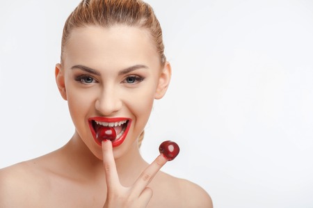 Attractive girl is holding two red cherries on her fingers. She is eating one cherry and laughing. Isolated on background and there is copy space in right sideの写真素材