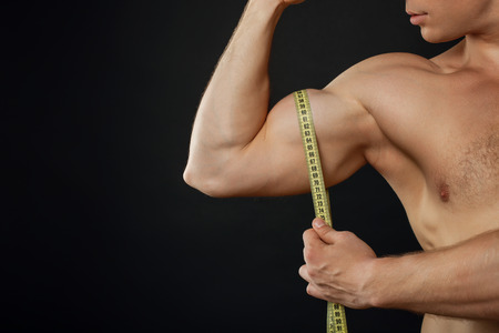 Close up of hand of bodybuilder measuring his bicep with meter. He is flexing. Isolated on black background and copy space in left sideの写真素材