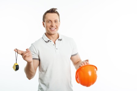 Handsome male worker is holding orange helmet and tape-measure in his hands. He is smiling and looking at the camera with joy. Isolated on background and copy space in right sideの写真素材