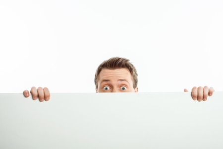 Cheerful guy is standing behind horizontal empty whiteboard. He is peeping through it with interest. Isolated on backgroundの写真素材