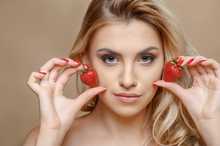 Beautiful blond girl is raising two strawberries to her cheeks. She is looking at the camera with temptation. Isolated on brown backgroundの写真素材