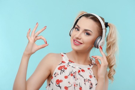 Cheerful woman is showing okay sign. She is listening to music from headphones. The lady is smiling and touching the equipment with enjoyment. Isolated on blue backgroundの写真素材