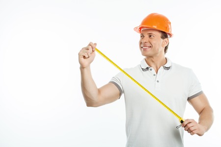 Handsome builder is holding tape-measure in his hands. He is looking aside and smiling. Isolated on background and copy space in left sideの写真素材